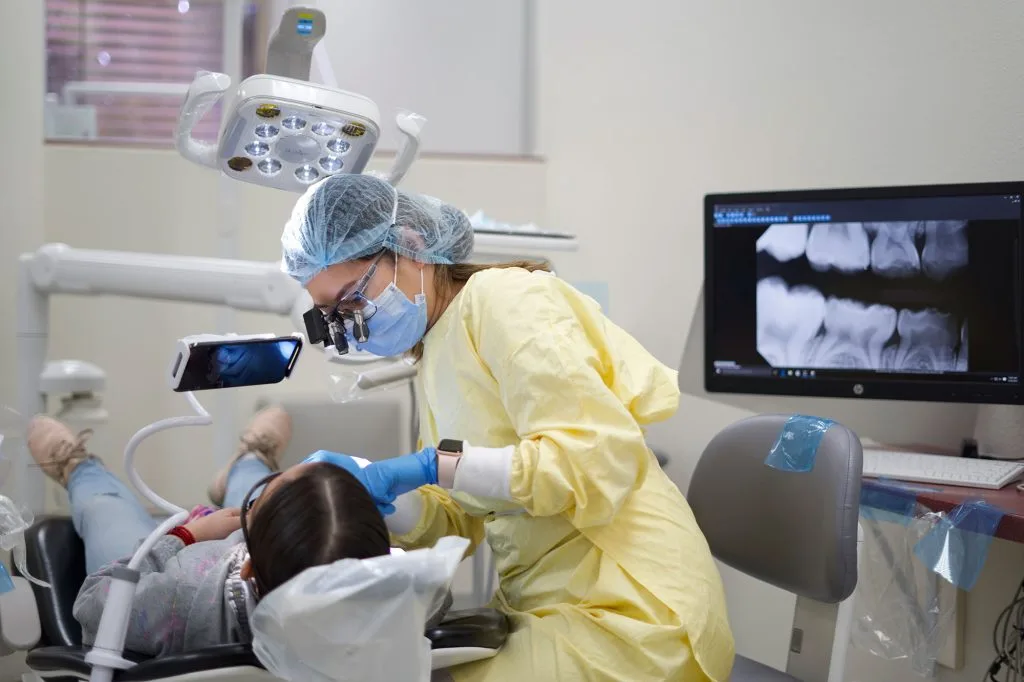 A dentist in protective gear treating a patient in a modern clinic, with dental X-rays visible on a screen - an example of the professional environment dental offices advertise to build trust.