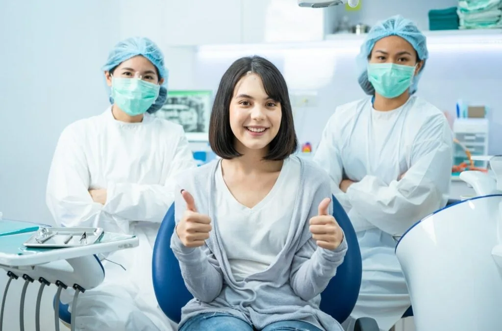 A young woman smiling and giving a thumbs up in a dental chair with two masked dentists in the background, highlighting the target audience of young adults who value modern, hygienic, and professional dental care.
